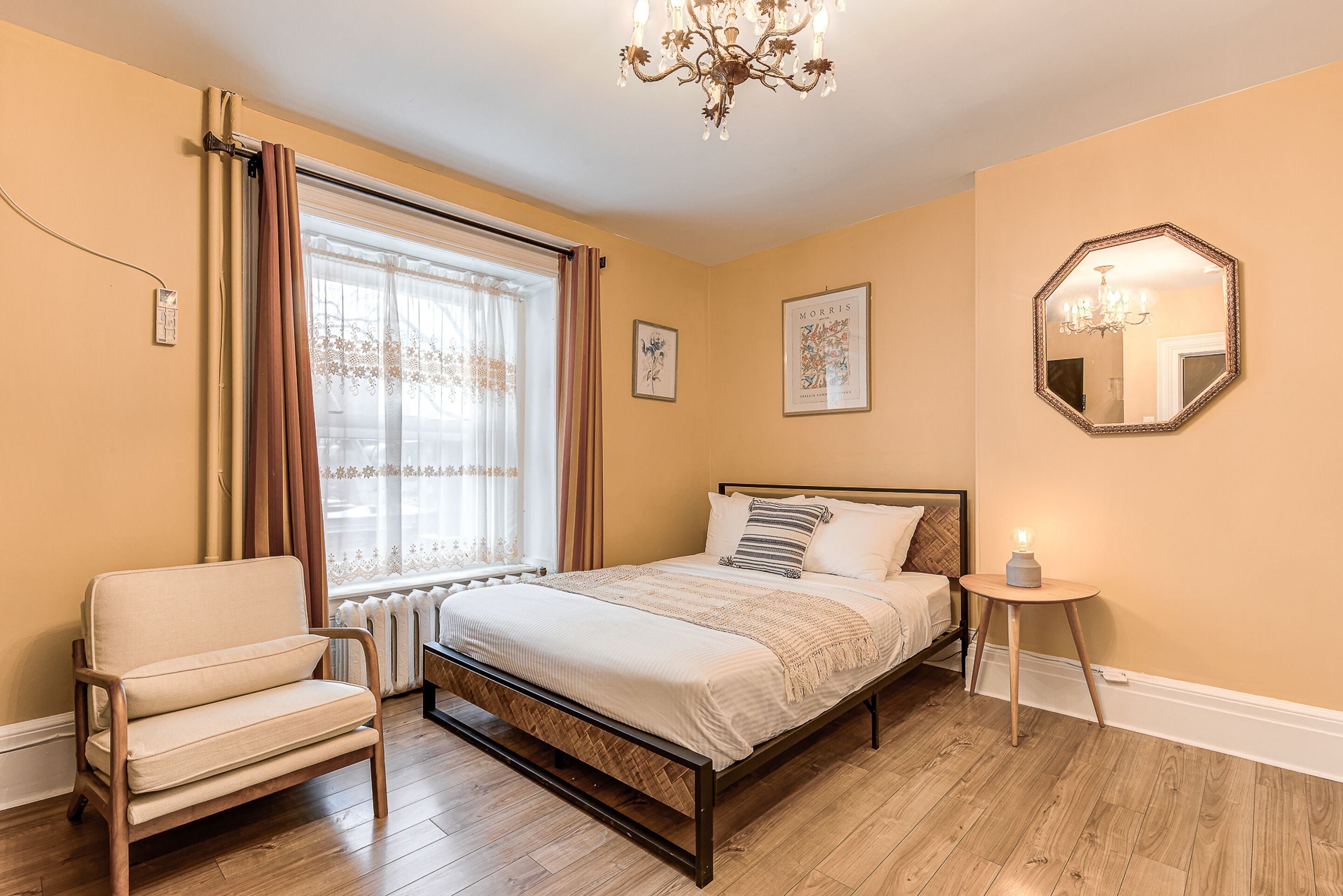 Une chambre à coucher comprenant un lit, une chaise, une table de chevet avec une lampe, une fenêtre avec des rideaux et un miroir sur le mur.