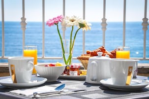 Desayuno con vista al mar, que incluye dos tazas de café, un vaso de jugo de naranja, un bol de fruta y un florero con flores.