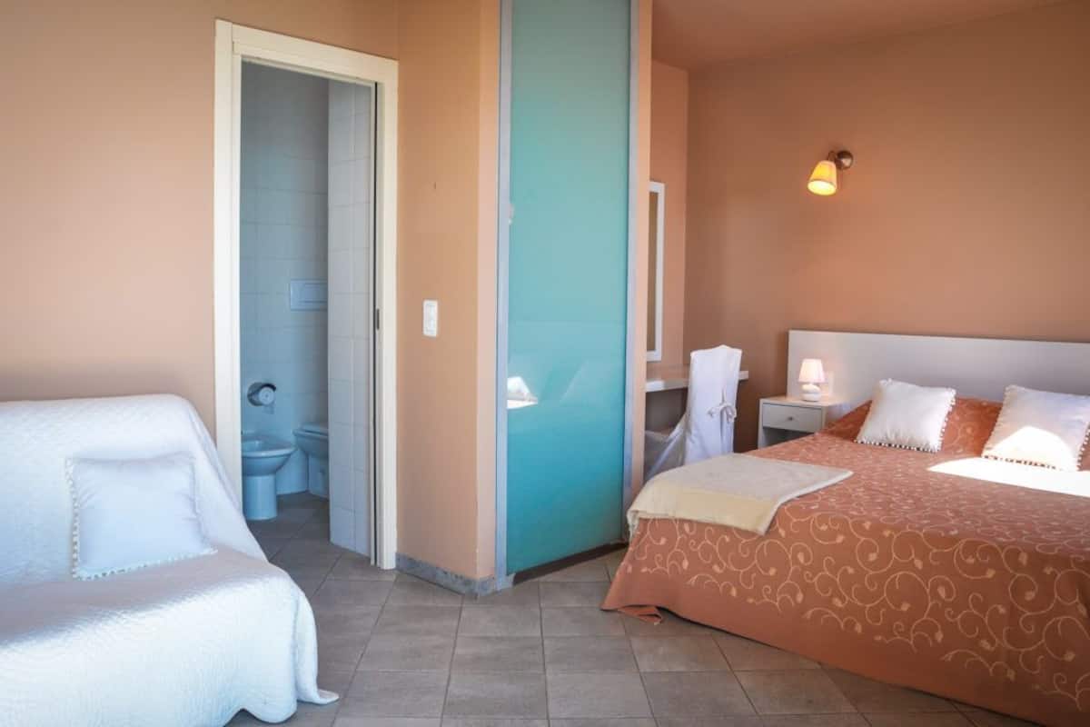 Ein Hotelzimmer mit einem Bett, einem Nachttisch mit Lampe, einem Badezimmer mit Toilette und Waschbecken sowie einer Tür zu einem weiteren Raum.
