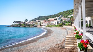 A coastal hotel with a pebble beach, steps leading to a covered area, and houses in the background.