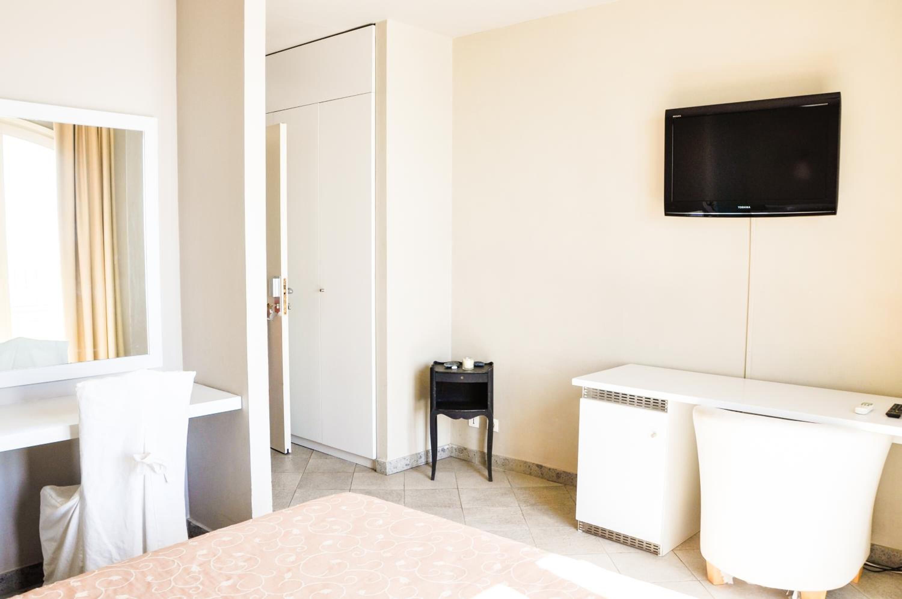 A hotel room with a bed, a desk, a chair, a TV mounted on the wall, and a door leading to another room.