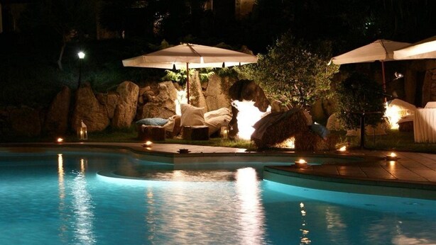 A nighttime swimming pool area with lounge chairs and umbrellas, surrounded by rocks and trees. - Funtana Abbas (Luras)