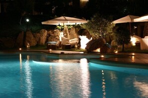 A nighttime swimming pool area with lounge chairs and umbrellas, surrounded by rocks and trees. - Funtana Abbas (Luras)