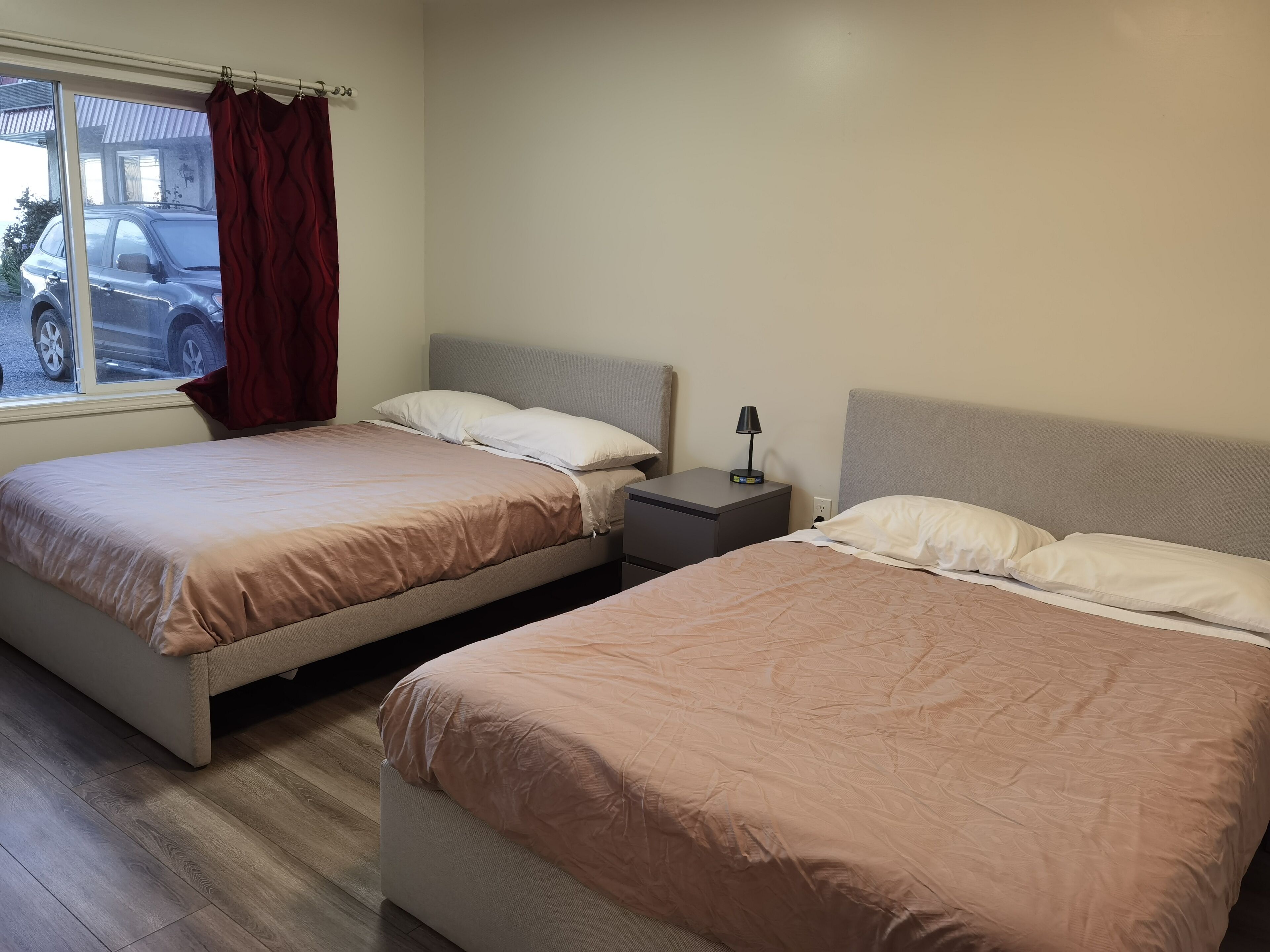 A hotel room with two beds, a nightstand with a lamp, and a window with a view of a parked car outside.
