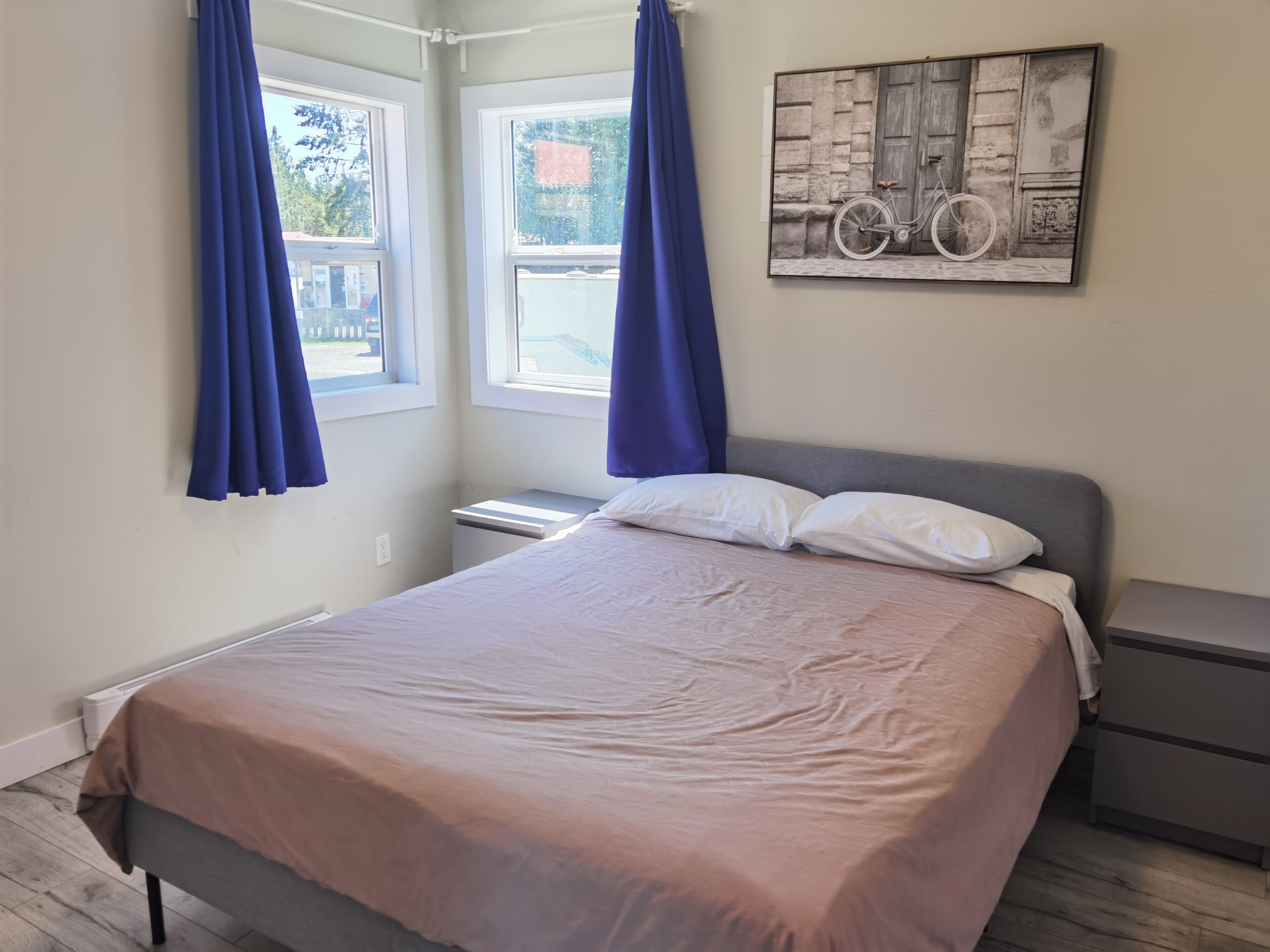 A neatly made bed with a grey headboard, a bedside table with a lamp, and a window with blue curtains.