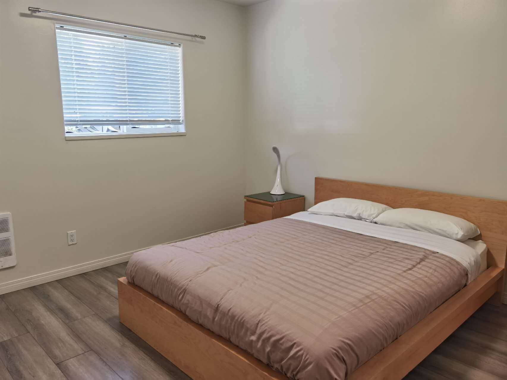 A bedroom with a bed, a nightstand, a bedside lamp, a window with blinds, and a wall-mounted heater.