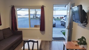 Una habitación con sofá, mesa, silla, televisor y vista a un estacionamiento con coches y una masa de agua.