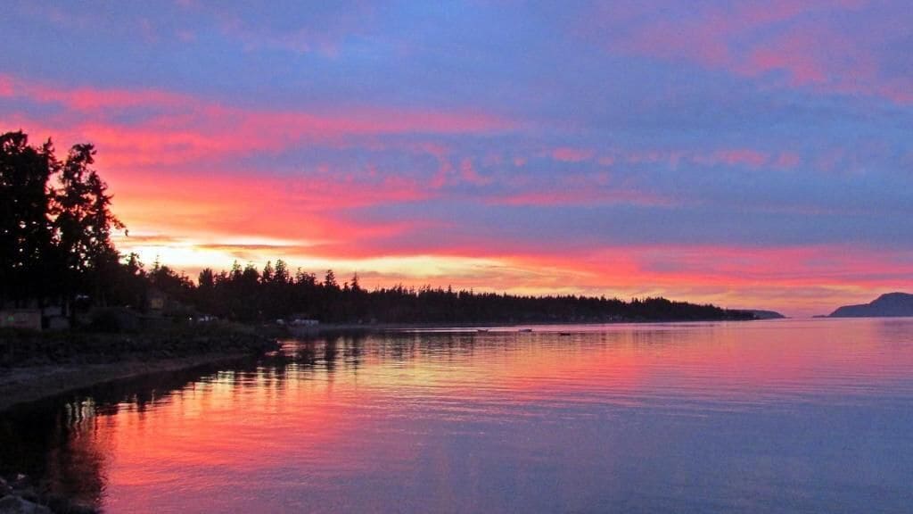 A calm body of water reflecting a sunset sky with a forested shoreline.