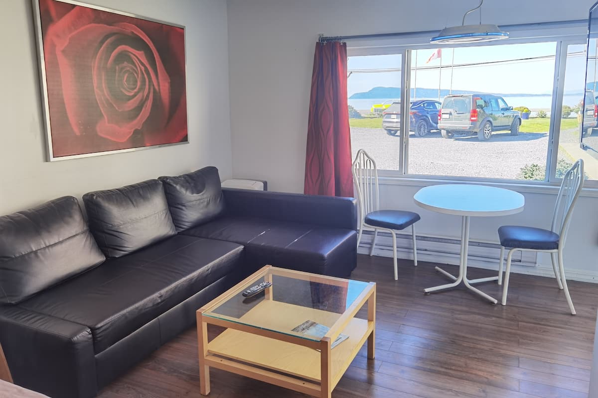 A modern room with a black leather sofa, a glass-top coffee table, and a view of parked cars and hills through a large window.