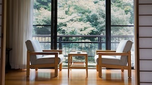Une pièce avec deux chaises en bois et une petite table, de grandes fenêtres donnant sur des arbres et des fleurs, et un plancher en bois.