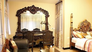 Una habitación de hotel con un espejo grande y ornamentado, una cama con cabecera decorativa, un escritorio de madera con una silla, un sofá y una mesita con una caja de pañuelos.