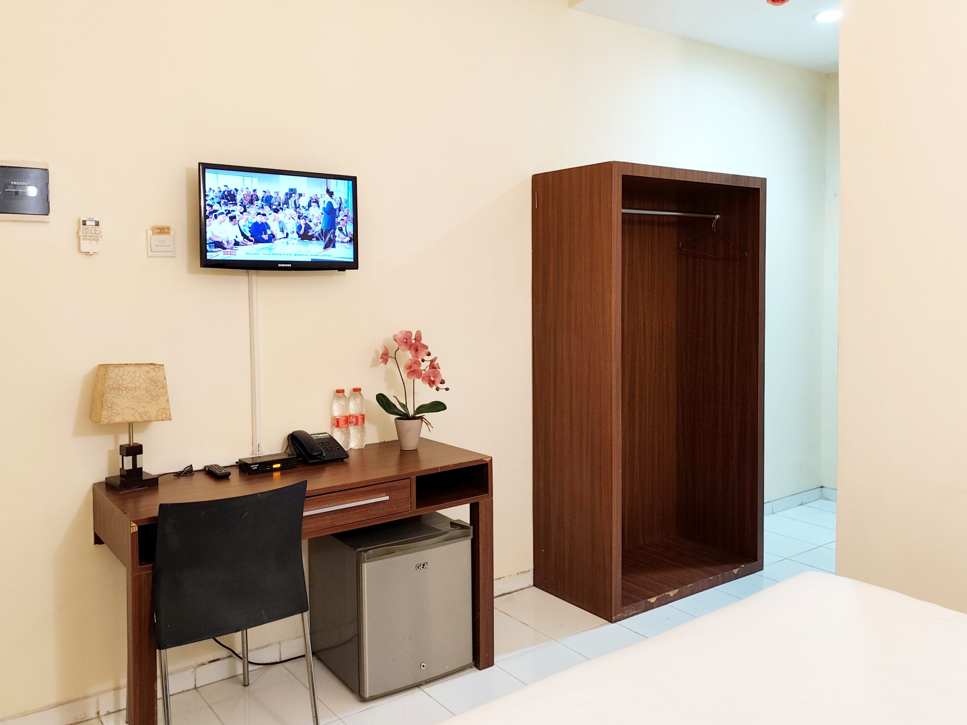 A hotel room with a wooden desk, a small fridge, a TV mounted on the wall, a lamp, a plant, and a wardrobe.