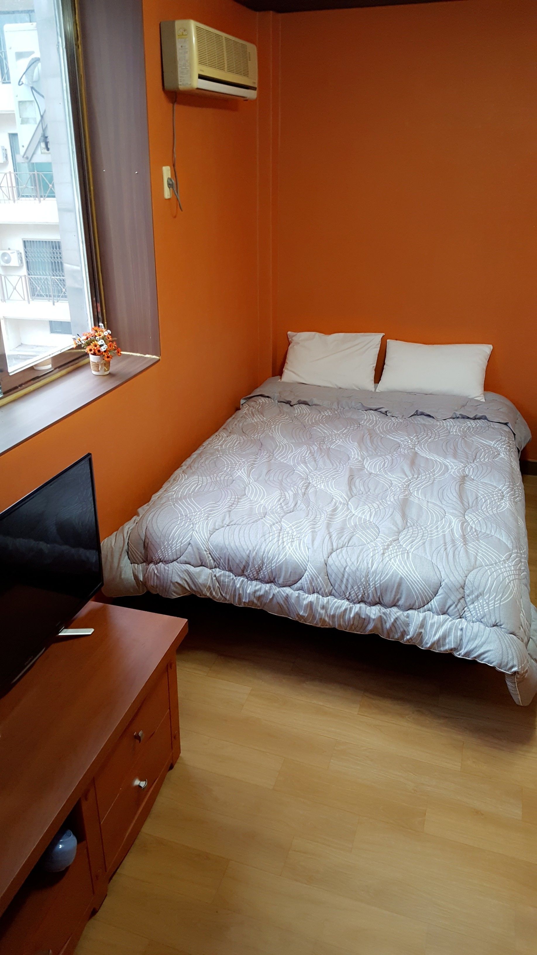 Et soveværelse med en orange væg, en seng med et mønstret dynebetræk, et træskrivebord med et fjernsyn og en aircondition på væggen.