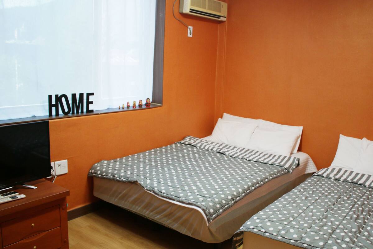 A room with two beds, a TV on a wooden cabinet, and a window with 'HOME' written on it.