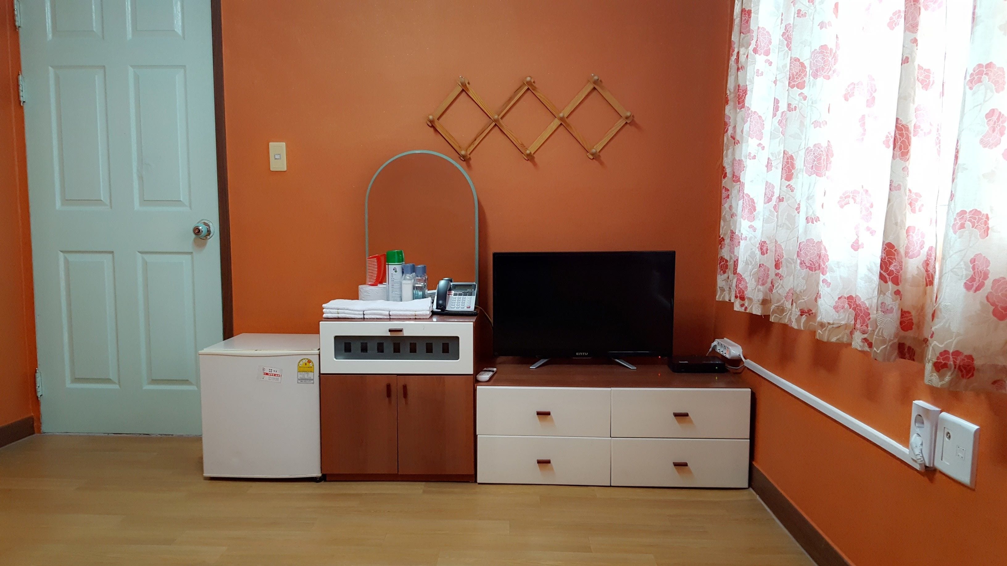 Een kamer met een witte deur, een oranje muur, een bureau met een flatscreen-tv, een telefoon en een kleine koelkast.