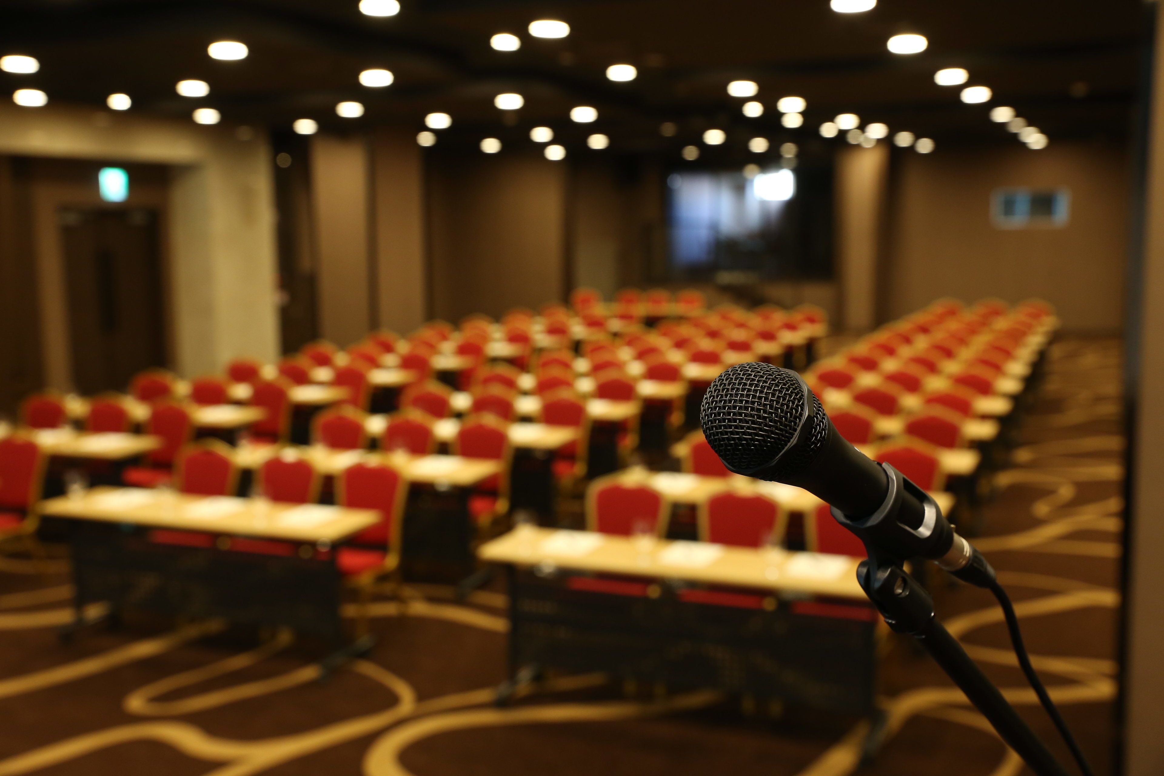 A conference room with rows of red chairs and tables, a microphone on a stand, and a carpeted floor.