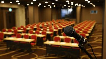 A conference room with rows of red chairs and tables, a microphone on a stand, and a carpeted floor.