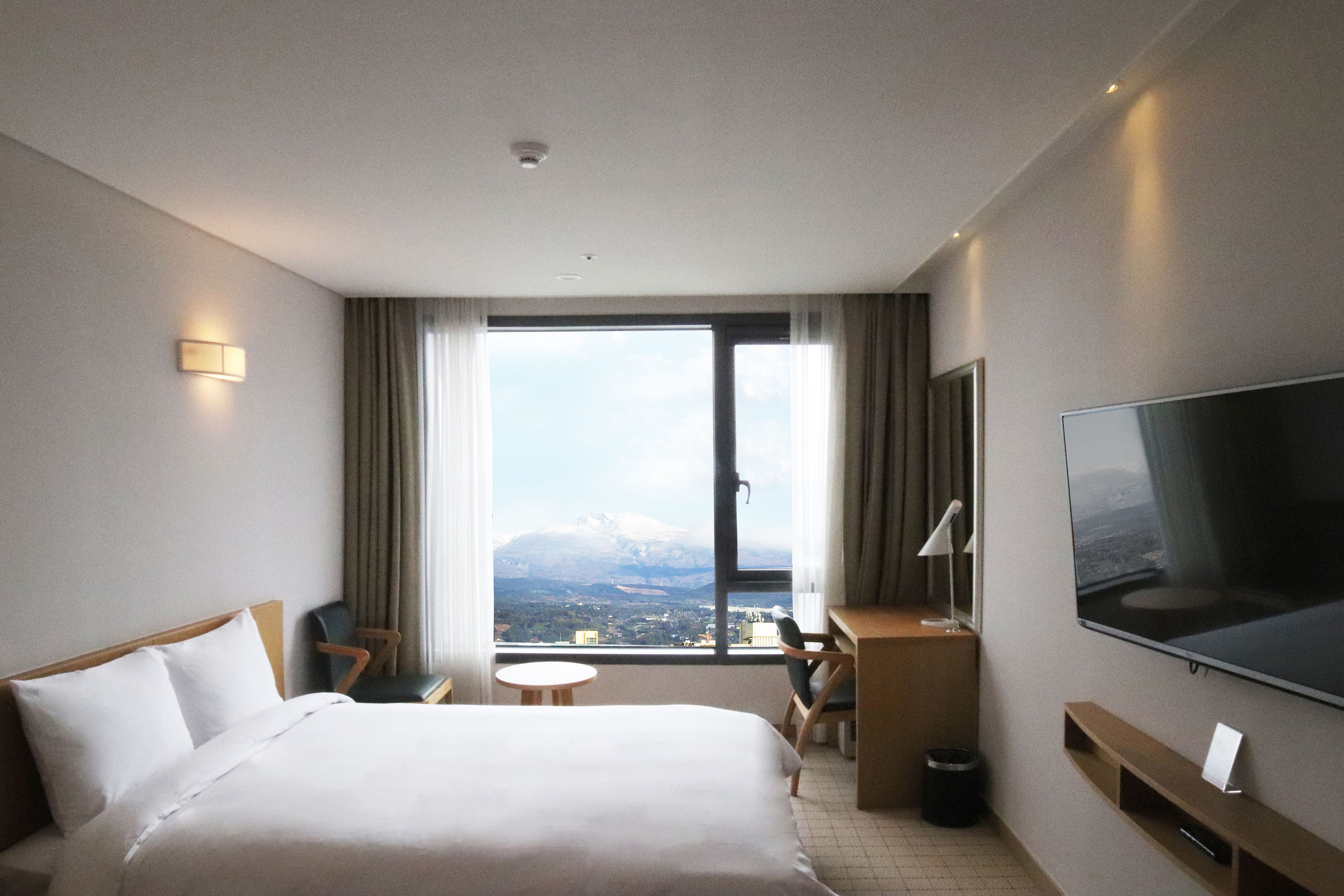 A hotel room with a bed, a desk, a chair, a TV, and a window with a mountain view.