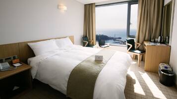 A hotel room with a large bed, a desk with a laptop, a chair, a trash bin, and a view of the sea and a bridge.