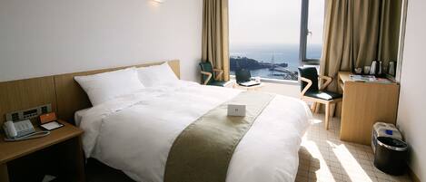 A hotel room with a large bed, a desk with a laptop, a chair, a trash bin, and a view of the sea and a bridge.