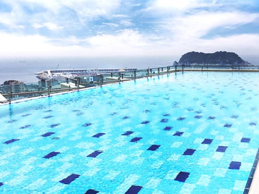 A rooftop infinity pool with clear blue water, a metal railing, and a view of the sea and distant buildings.