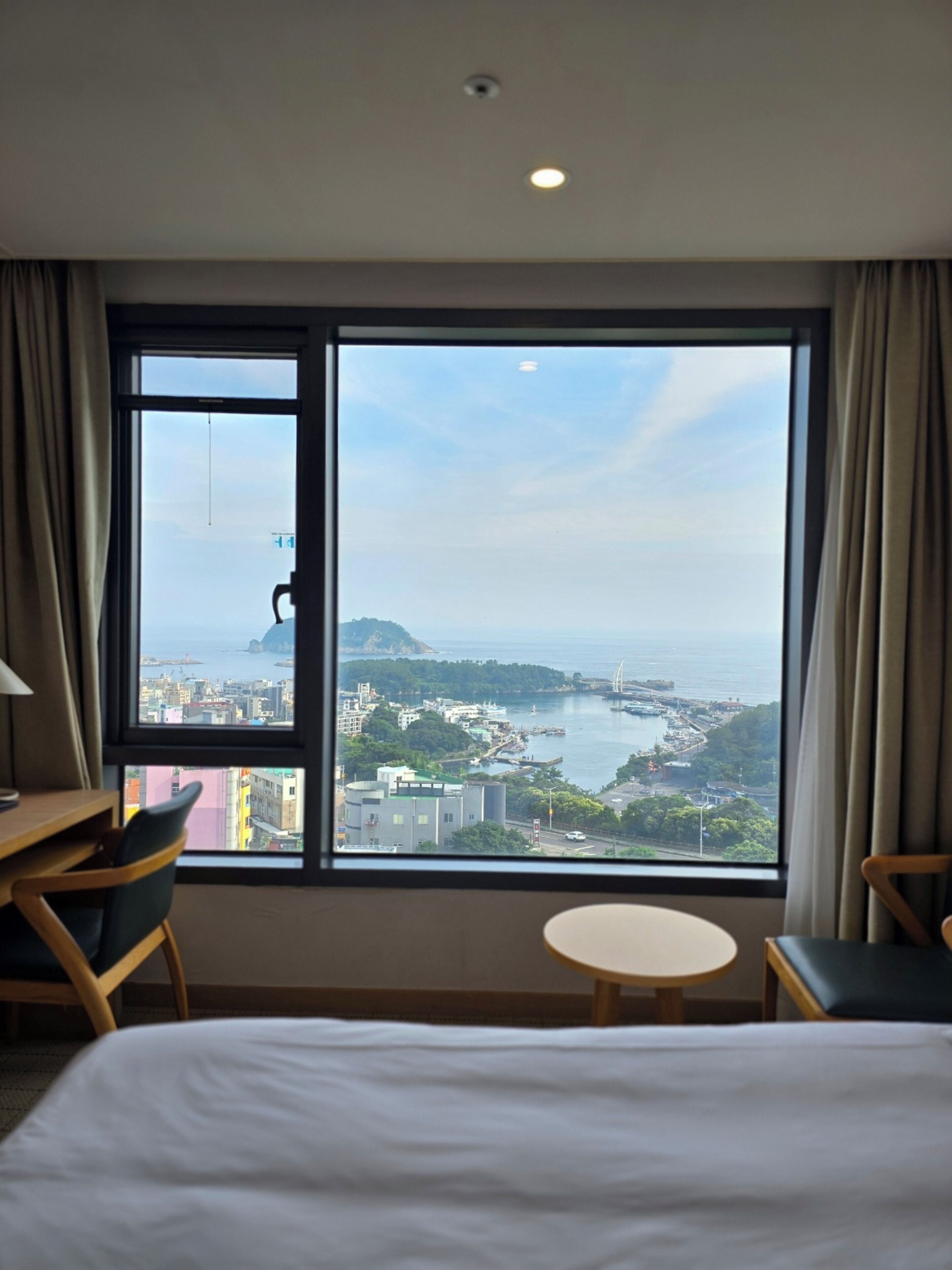 Quarto de hotel com uma janela ampla que oferece vista para a cidade costeira, incluindo uma ponte e o porto. Há uma cama, duas cadeiras e uma pequena mesa.