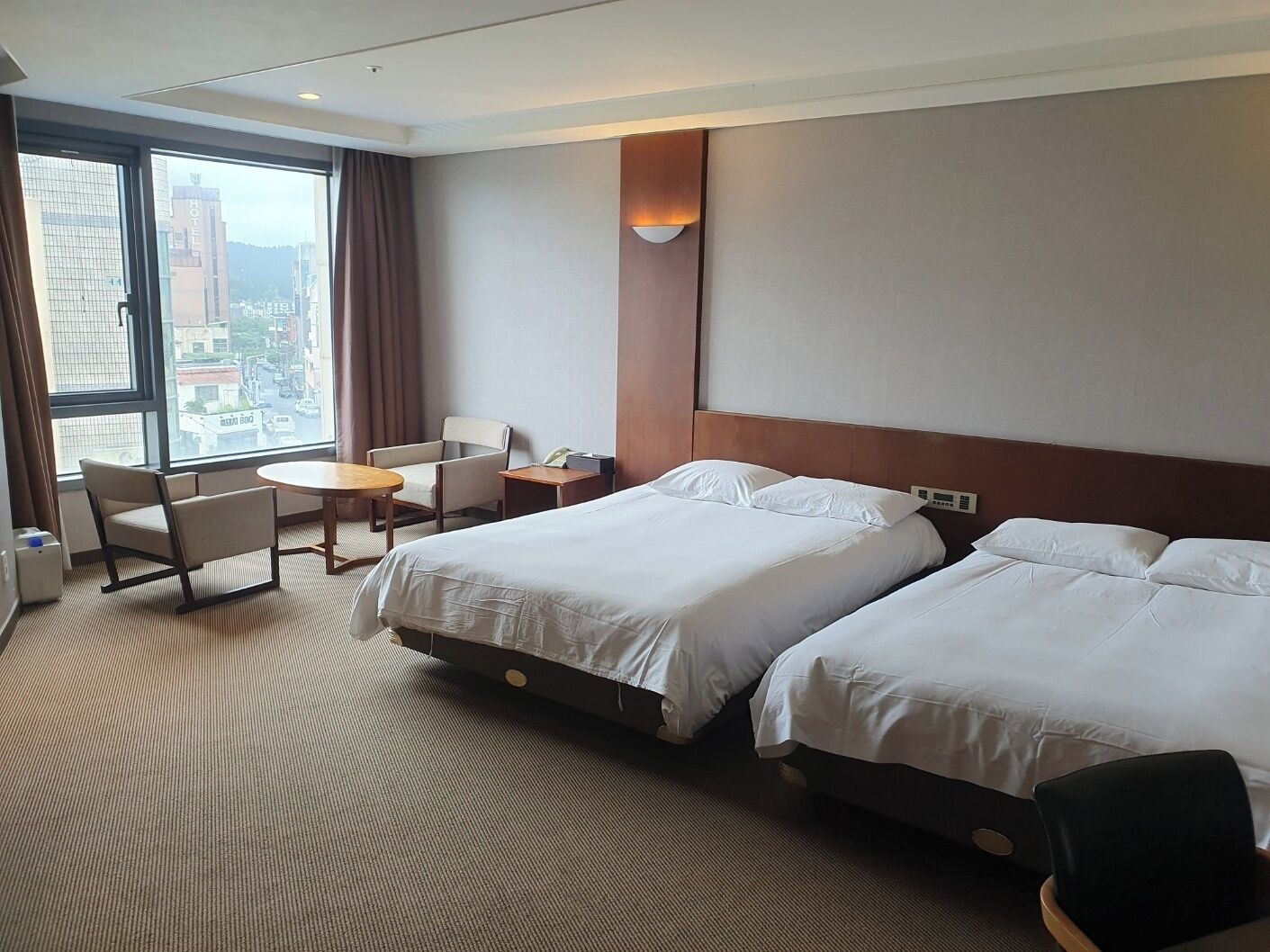 A hotel room with two beds, a desk, a chair, and a large window offering a city view.