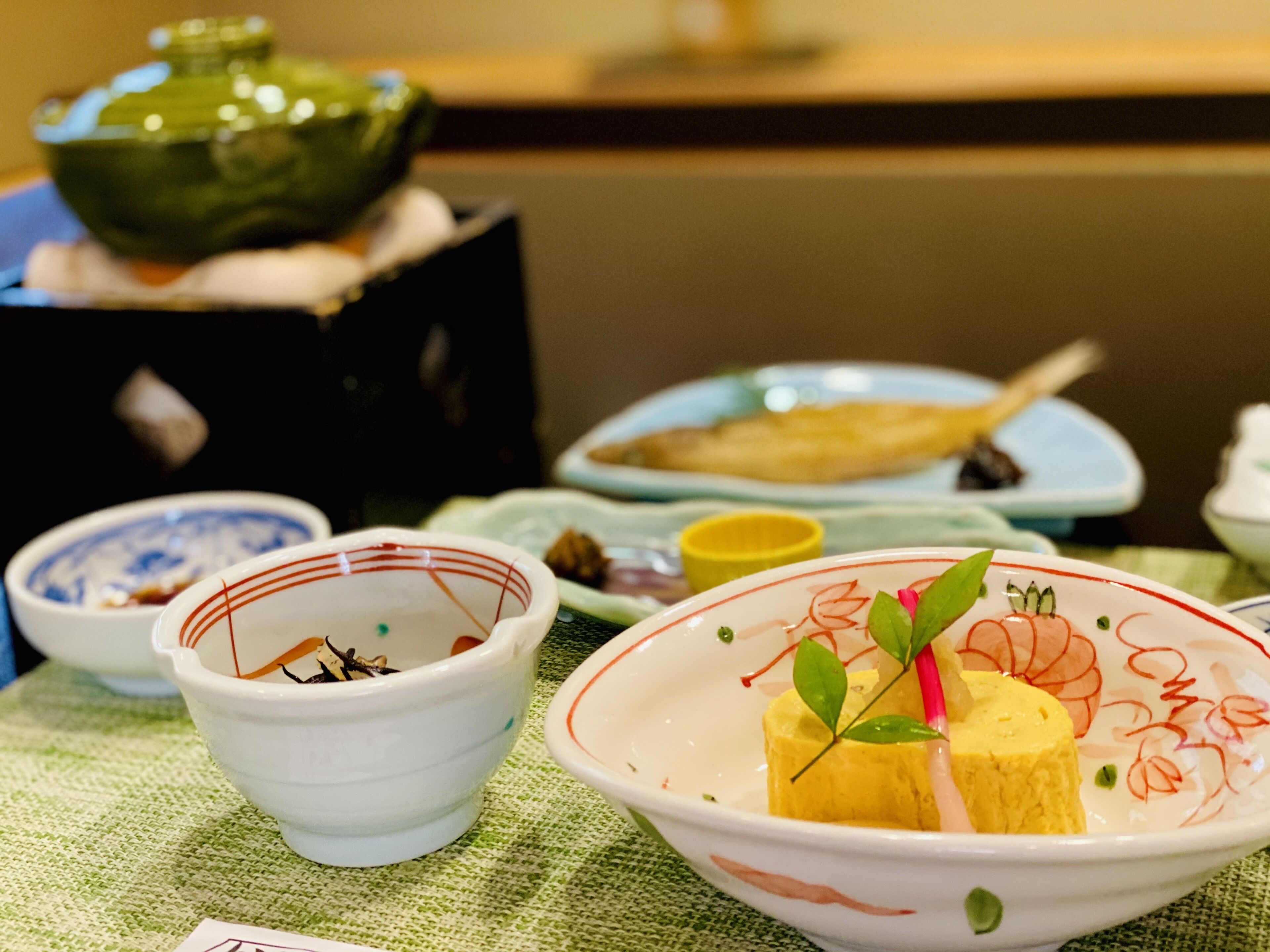 A table set with various dishes, including a bowl with a yellow item and green leaves, and a green teapot in the background.