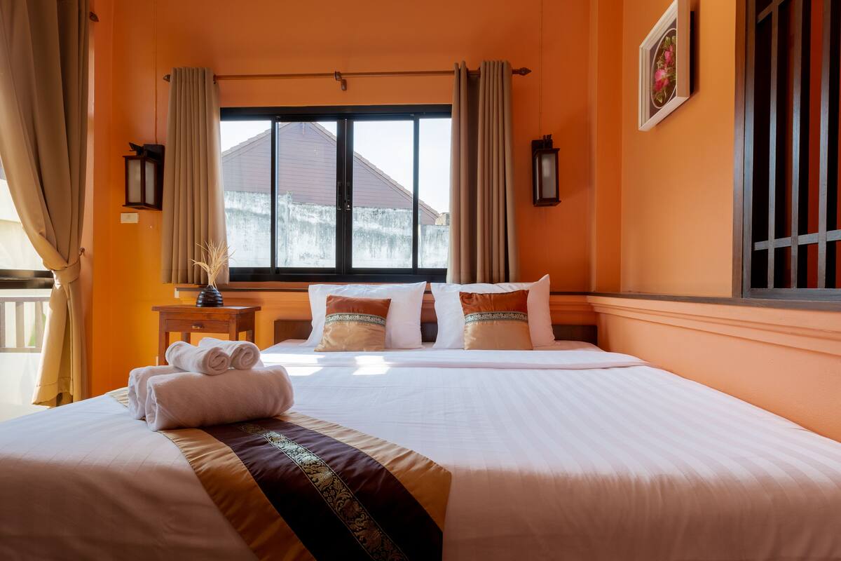 A hotel room with a large bed, beige curtains, a window with a view of a building, and a small table with a vase.