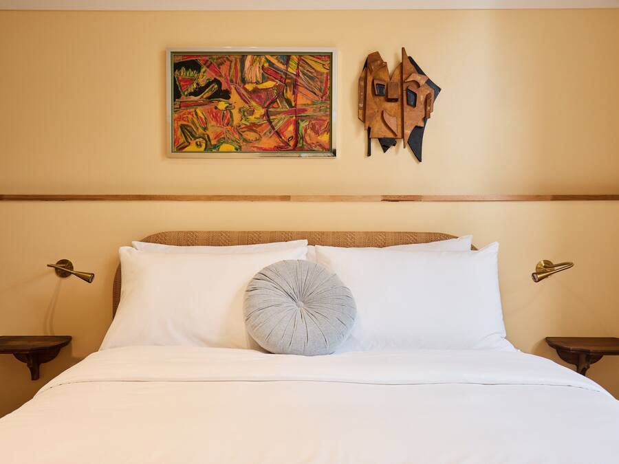 Une chambre d’hôtel avec un lit, du linge de lit blanc, un tableau abstrait aux couleurs vives et une sculpture en bois.