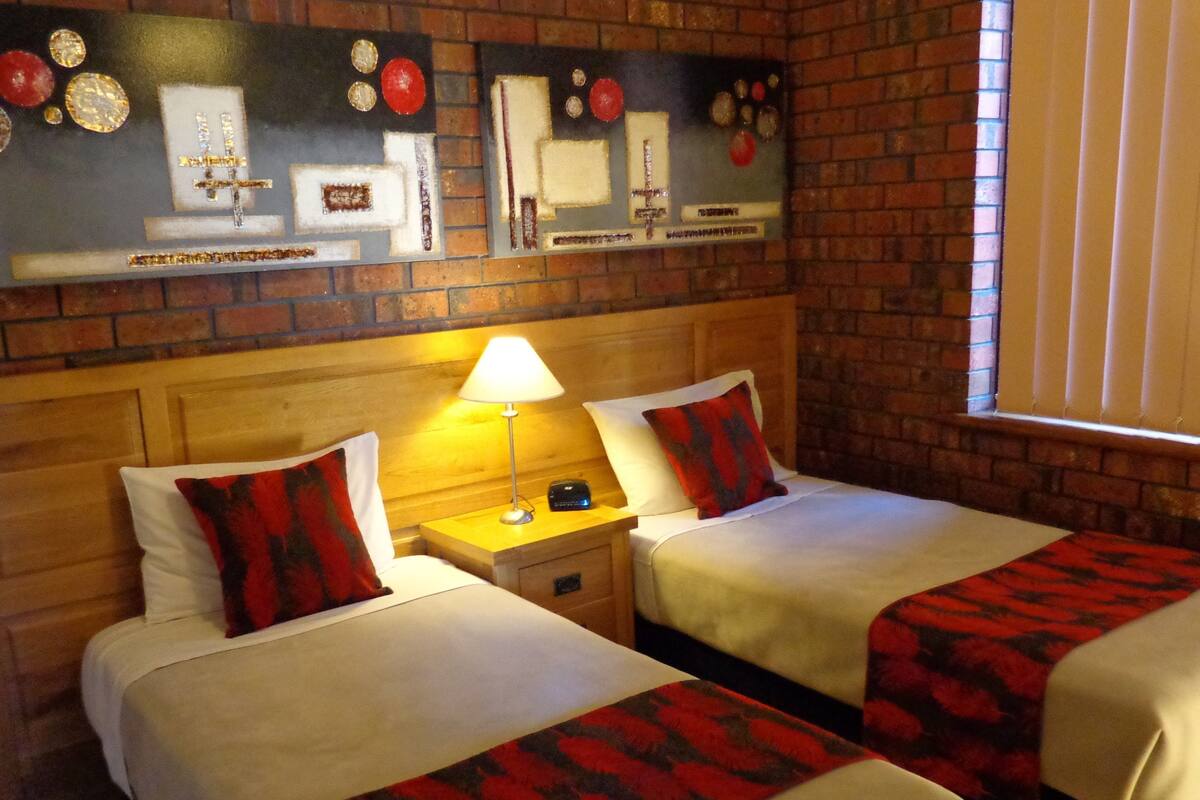 A hotel room with two beds, a wooden headboard, a nightstand with a lamp, and a decorative wall piece above the beds.