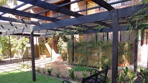 A covered patio area with a brick pathway, a bench, and potted plants. - Glenelg Gateway Apartments (Glenelg)