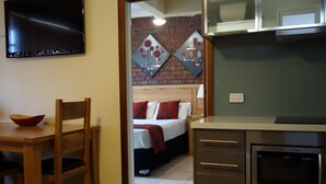 A compact hotel room with a wooden dining table, a bed with red and white bedding, a microwave, and a TV mounted on the wall. - Glenelg Gateway Apartments (Glenelg)