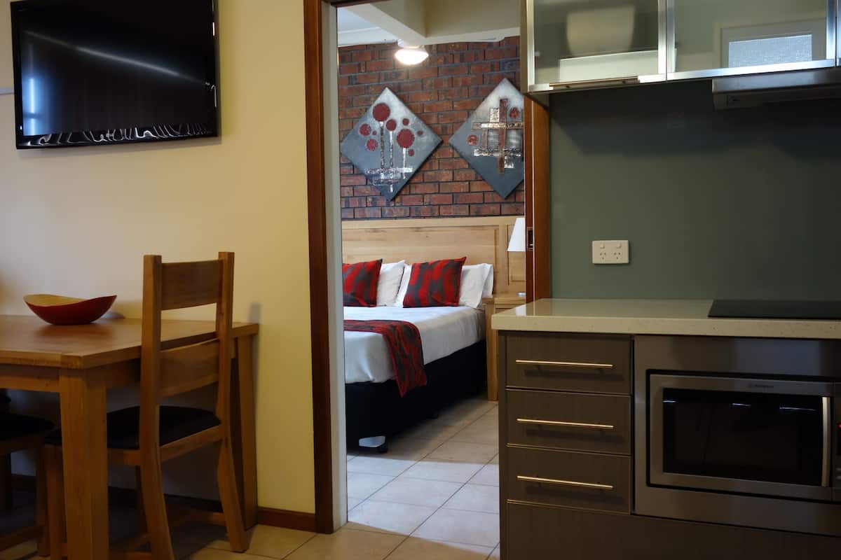 A compact hotel room with a wooden dining table, a bed with red and white bedding, a microwave, and a TV mounted on the wall.