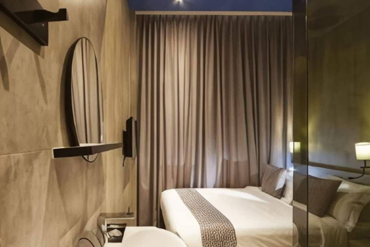 A modern hotel room with a large bed, a bedside table, a mirror, and a curtain.