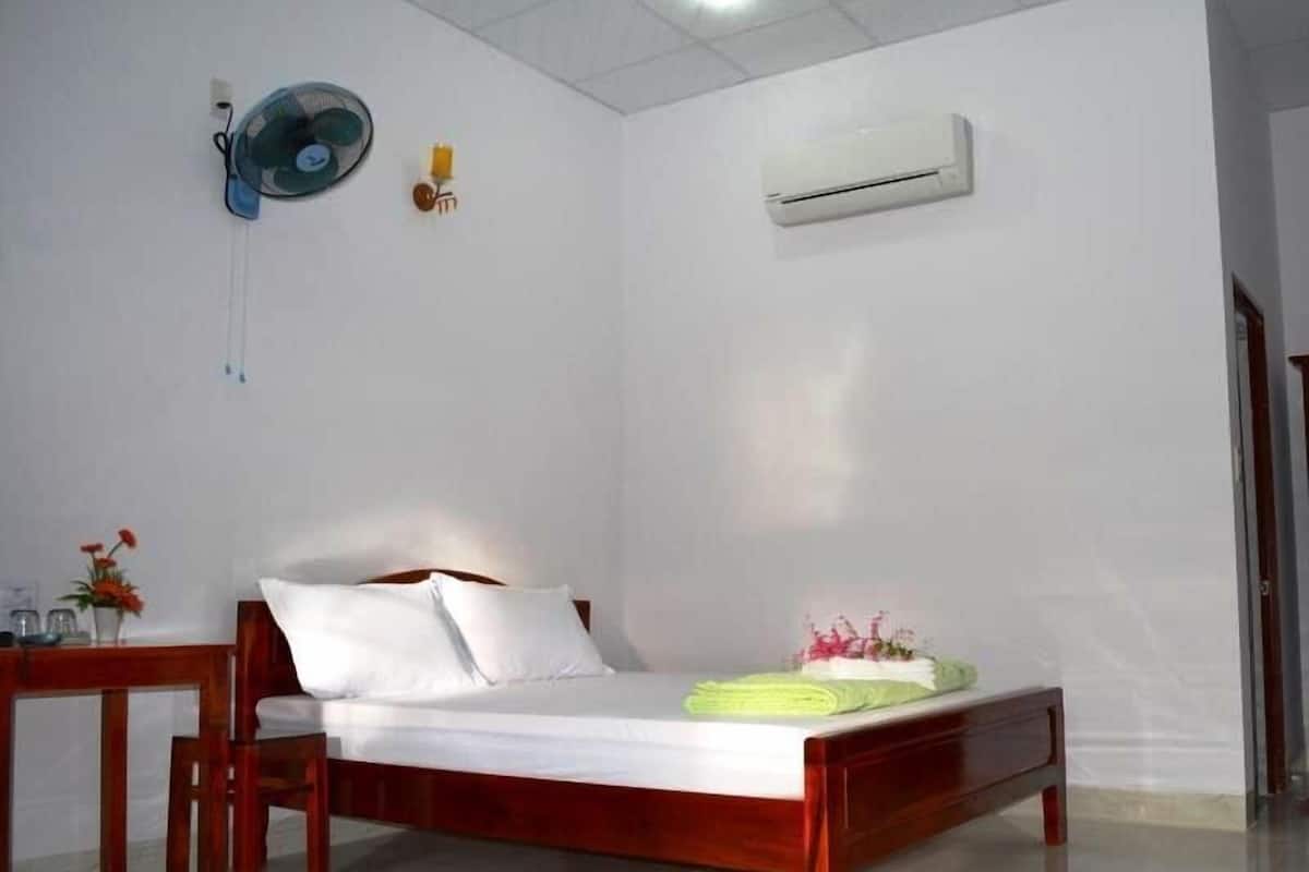 A bedroom with a wooden bed, white walls, a ceiling fan, an air conditioner, and a small table with a vase of flowers.