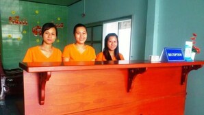 A reception desk with three individuals seated behind it, a blue sign labeled "RECEPTION", and a wall with floral decorations. - Diem Lien Guesthouse (Mui Ne)