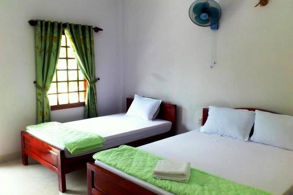 A room with two single beds, a window with green curtains, a wall-mounted fan, and a small wall decoration.