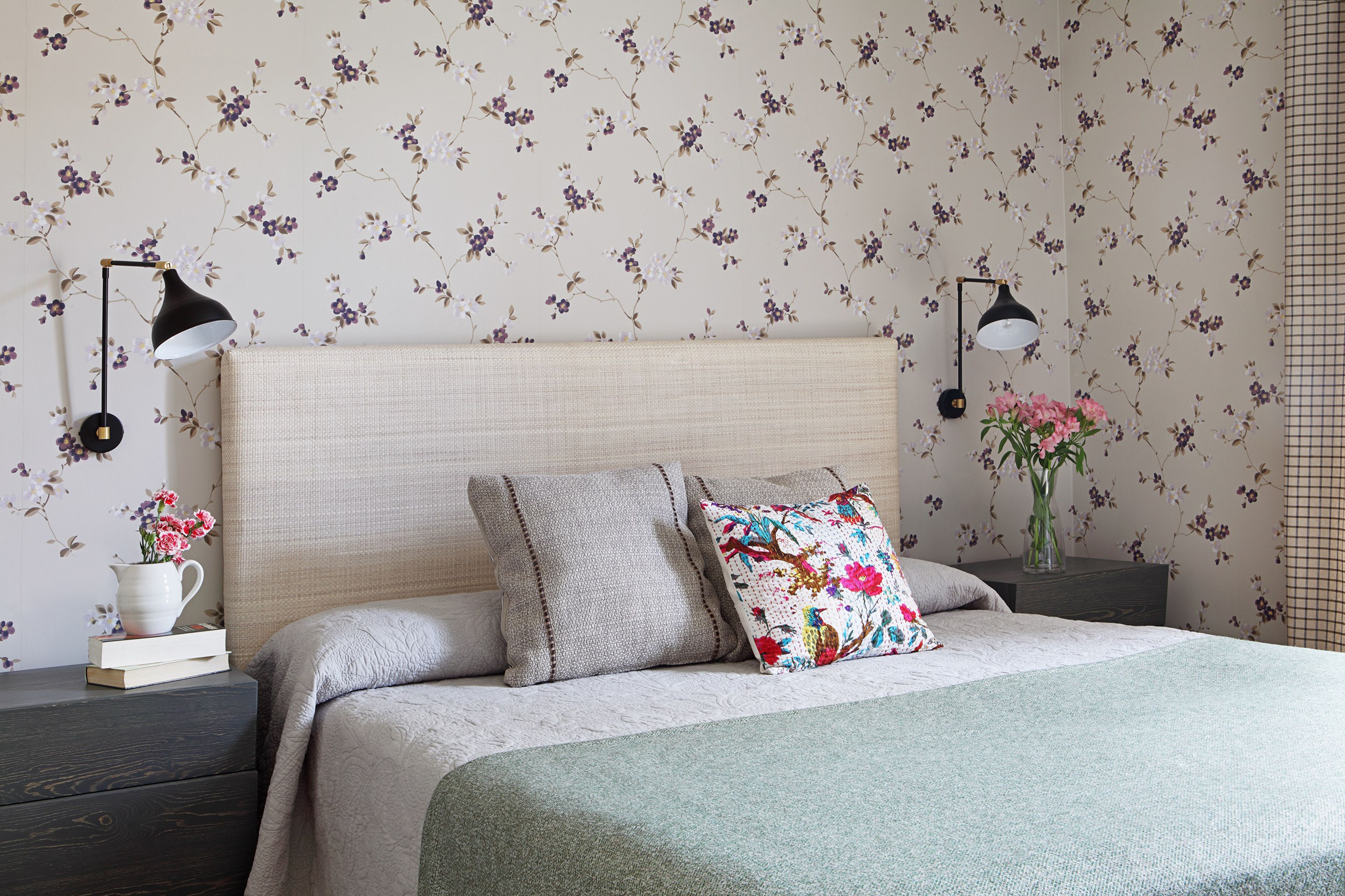 A neatly made bed with a floral-patterned headboard, a bedside table with a lamp, and a vase with flowers.