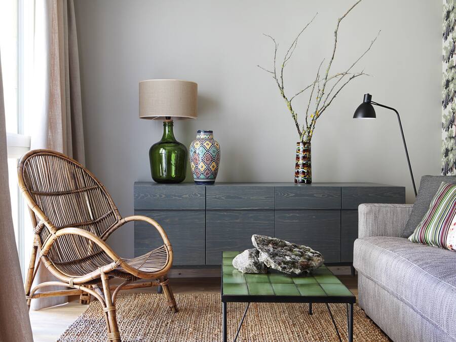 Un salón con una silla de mimbre, un sofá, una mesa de centro verde, un aparador con objetos decorativos y una lámpara.