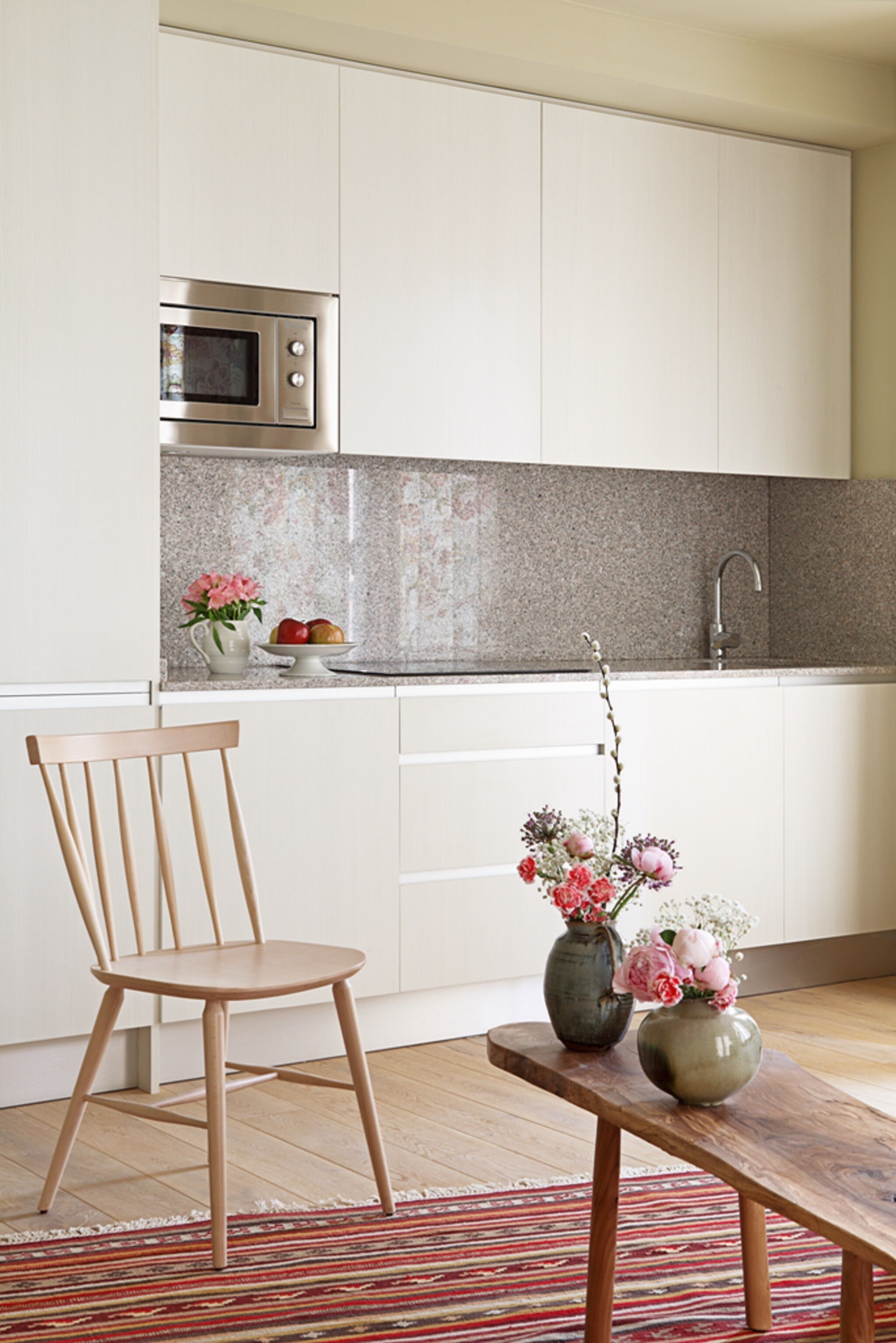 A kitchen with white cabinets, a stainless steel microwave, a wooden chair, a table with two vases of flowers, and a striped rug.