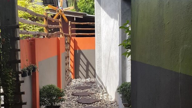 Un estrecho sendero con una valla de madera y una puerta, que conduce a un edificio con tejado de baldosas y un pequeño árbol.