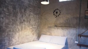 Una cama individual con ropa blanca en una habitación con pared de hormigón y un reloj.