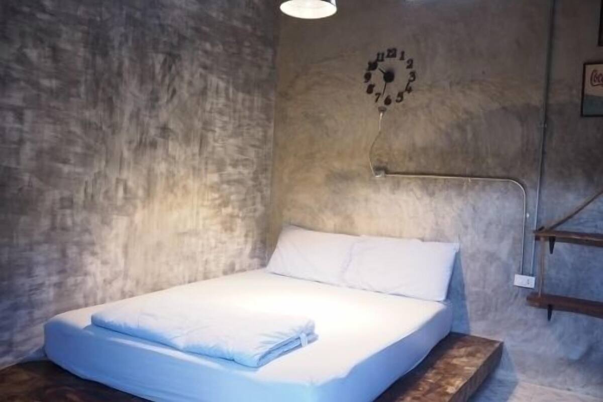 Una cama individual con ropa blanca en una habitación con pared de hormigón y un reloj.