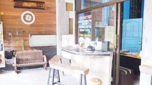 Una zona de bar con taburetes y una barra, una pared de madera con un reloj, y una puerta azul al fondo.
