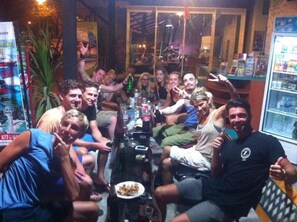 A group of people gathered around a table with drinks and snacks, in a room with a glass wall and a refrigerator in the background.
