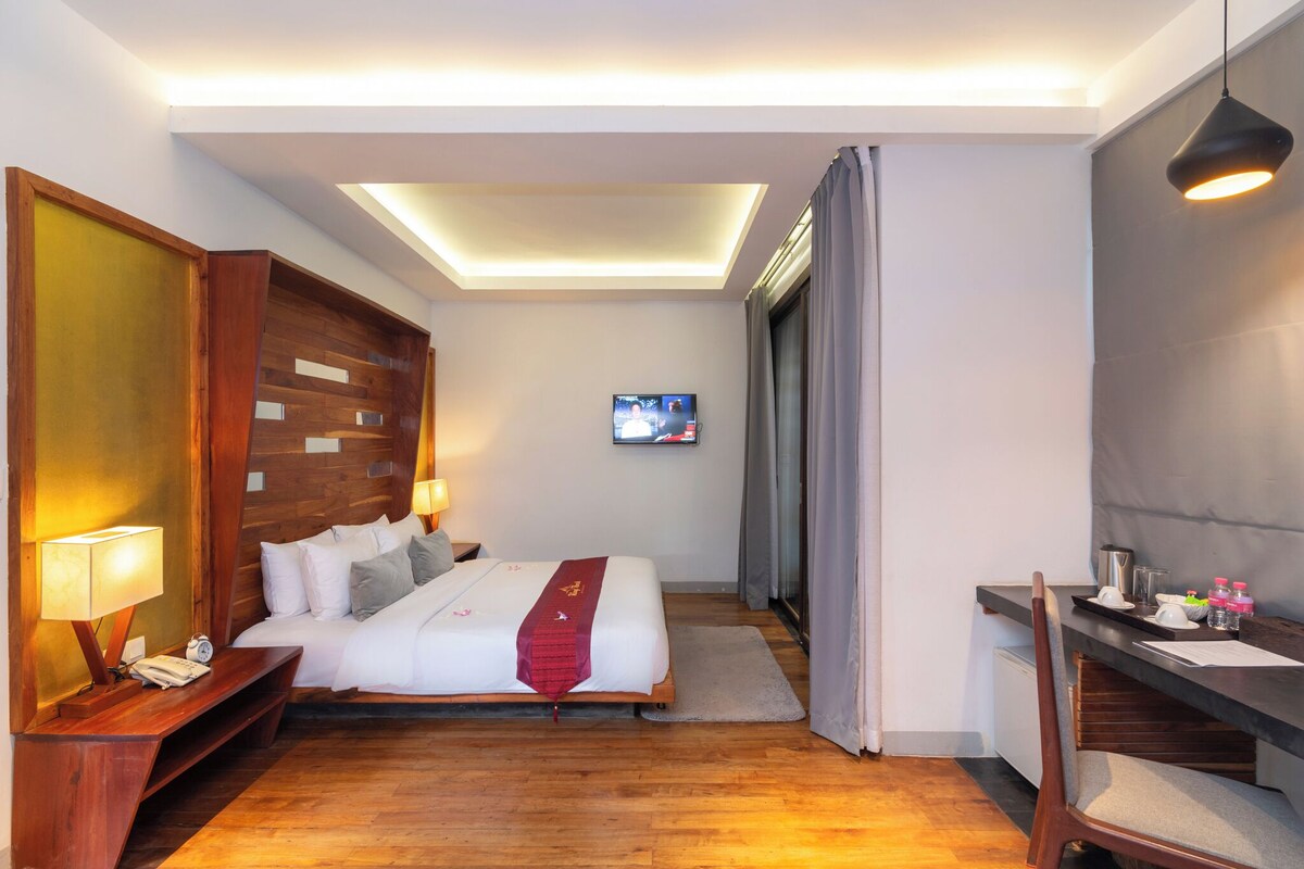 A hotel room with a bed, bedside table, TV, desk, chair, and a small kitchenette.