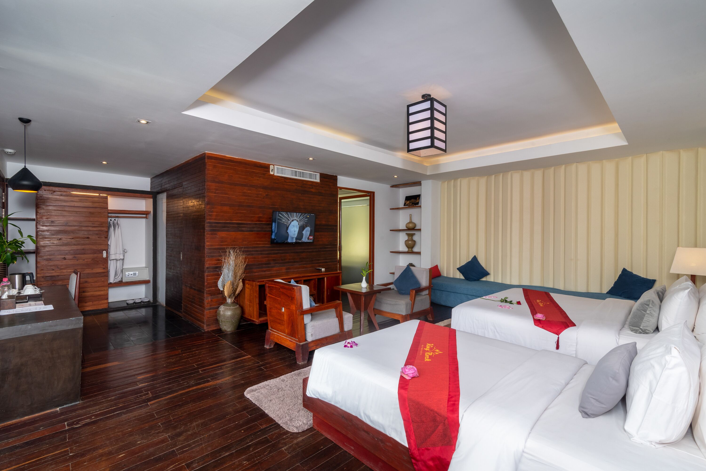 Ein Hotelzimmer mit einem hölzernen Empfangstresen, einem Fernseher, einem Bett mit roter und weißer Bettdecke, einem blauen Sofa und einem großen Fenster.
