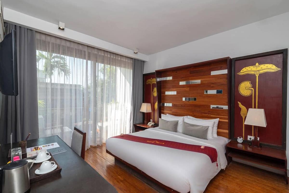 A hotel room with a large bed, a wooden headboard, a desk with a coffee maker, and a view of palm trees through the window.