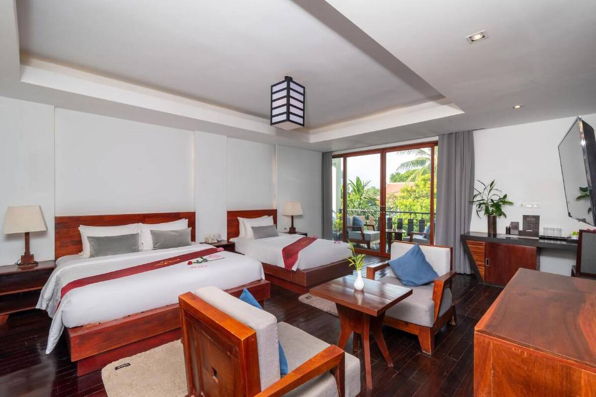 A hotel room with two beds, a sofa, a desk, and a balcony with a view of greenery.